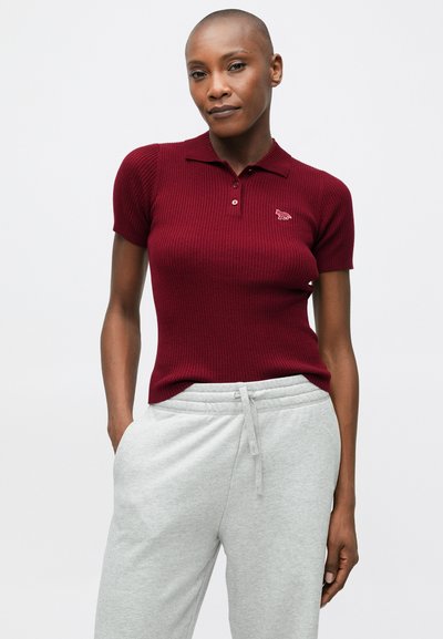 Sieviete ar īsām piedurknēm burgundijas krāsas ribota polo krekla un gaiši pelēkiem sporta šortiem, stāvot ar vienu roku kabatā.