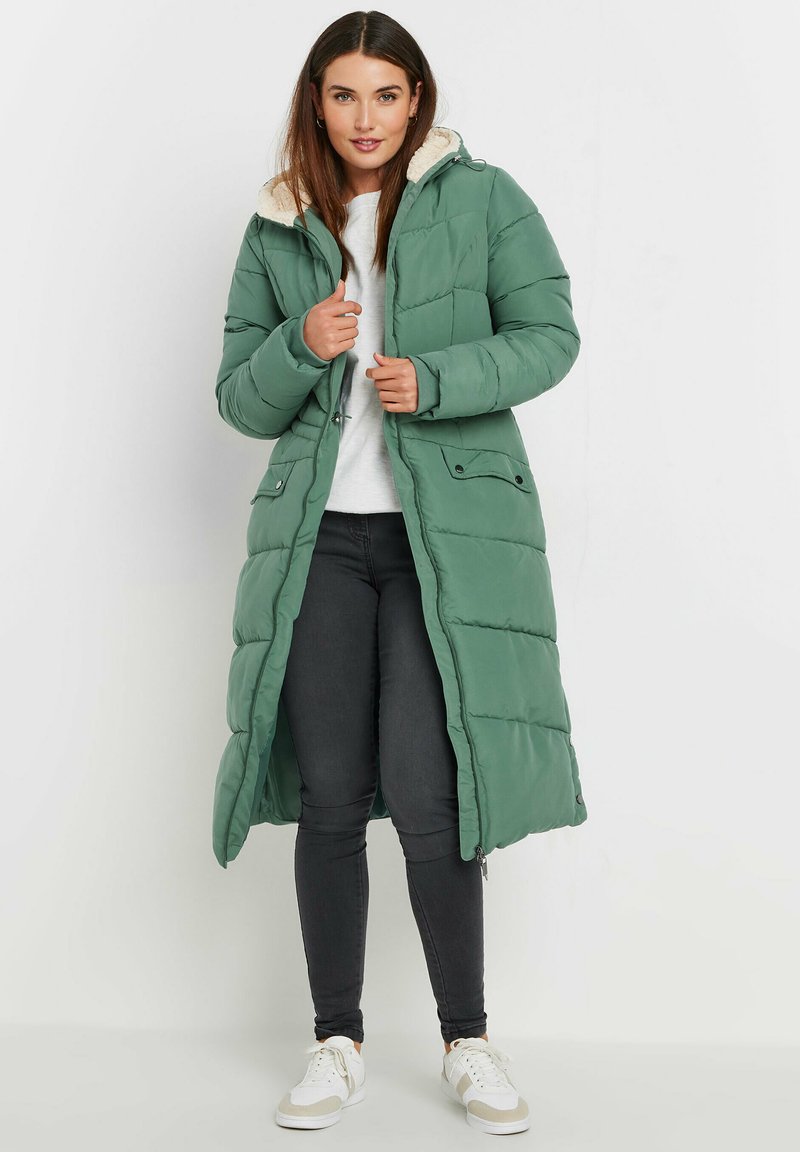 Lång, grön pufferjacka med foder av päls, tryckknappsstängningar och fickor. Matchad med grå byxor och vita sneakers.