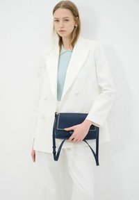 Marineblauwe leren handtas met witte stiksels en een afneembare schouderband. Model draagt een witte blazer en een geribde lichtblauwe top.