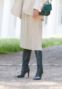 Femme portant une jupe plissée beige, un manteau beige, des bottes à talons noires et tenant un sac à main en cuir vert foncé, à l'extérieur sur un chemin en gravier.