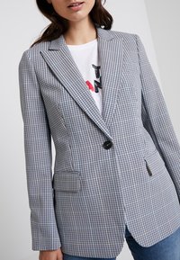 Blazer a scacchi in blu chiaro e nero, con un solo bottone, rever a punta e due tasche frontali. Indossato sopra una t-shirt grafica.