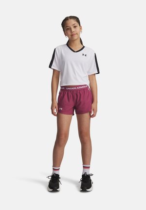 Noor tüdruk seisab, kandes valget Under Armour lühikest topi, burgundi värvi Under Armour lühikesi pükse, triibulisi sokke ja musti spordijalatsid.