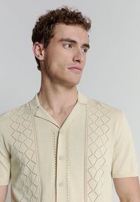 Lichtbeige polo-shirt met korte mouwen en open kraag, met een gestructureerd ruitpatroon en knoopsluiting aan de voorkant. Glad breigoed.