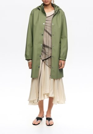 Frau trägt einen kniehohen olivgrünen Regenmantel über einem beigen, mehrlagigen Kleid mit schwarzen abstrakten Linien und schwarzen Sandalen mit offenen Zehen und Absatz.