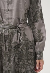 Sukienka z satyny w stonowanym szarym kolorze z zielonym wzorem paisley. Posiada zapięcie na guziki, długie rękawy i związany w talii pasek. Gładka tekstura.