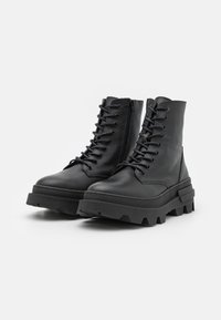Paire de bottes de combat en cuir noir à lacets, avec semelles en caoutchouc épaisses et zips latéraux, sur un fond blanc uni.