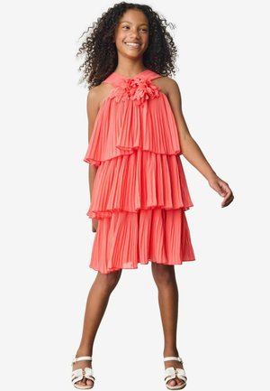 Robe plissée corail avec des niveaux superposés et un accent floral au niveau du col. Texture lisse, design sans manches, associée à des sandales blanches.
