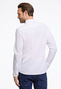 Camicia bianca in lino a maniche lunghe con colletto a punta, caratterizzata da una piega sul retro e bottoni dorati a contrasto sui polsini.