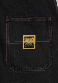 Poche de salopette en denim noir avec une étiquette rectangulaire dorée présentant un motif graphique. Couture contrastée en orange.