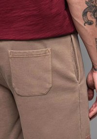Un pantalon de survêtement beige présente une seule poche plaquée à l'arrière. Le tissu semble doux avec une texture lisse et une coupe décontractée.