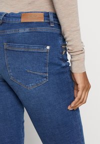 Gros plan d'une personne portant un jean en denim bleu avec une poche arrière et une étiquette en cuir marron sur la ceinture, associée à un haut beige à manches longues.