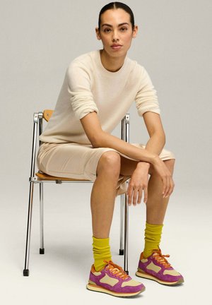 Les baskets présentent une tige violette et beige avec des lacets orange, assorties à des chaussettes côtelées jaunes vives ; le modèle porte un pull crème et une jupe beige.
