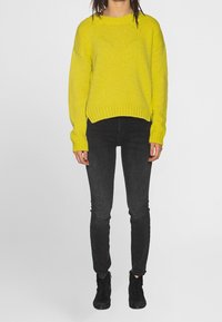 Jasně žlutý pletený svetr s žebrovaným lemem a manžetami, doplněný černými úzkými džíny a černými kotníkovými botami.