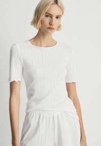 Top bianco in cotone a costine con maniche corte a scallop, caratterizzato da strisce verticali di tessuto e scollatura rotonda. Design morbido e aderente.