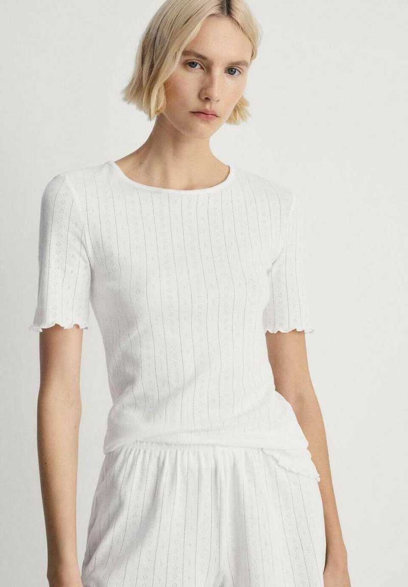 Top bianco in cotone a costine con maniche corte a scallop, caratterizzato da strisce verticali di tessuto e scollatura rotonda. Design morbido e aderente.