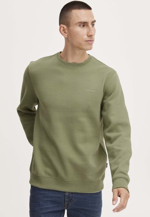 Olive roheline kapuutsiga särk, millel on ümar kaelus ja soonikkrae. Valmistatud pehmest kangast, millel on rinnal diskreetne brändi logo.