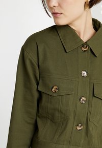 Veste en shirt de couleur olive, fabriquée à partir d'un tissu robuste, dotée d'un col pointu, de poches frontales et de grands boutons marron.