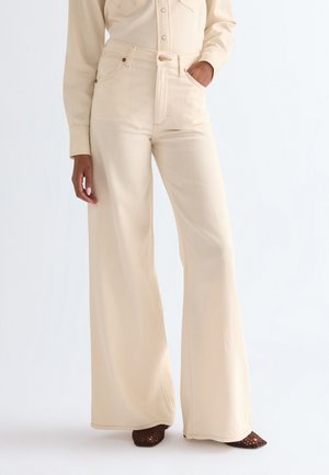 Vrouw draagt hoge taille, wijde beige broek en bijpassend overhemd, met één hand in de zak en bruine geweven schoenen.