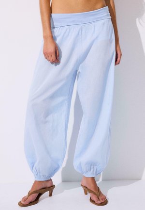 Žena v širokých svetlomodrých nohaviciach s elastickými manžetami a hnedých sandáloch na podpätku stojí na bielom pozadí.