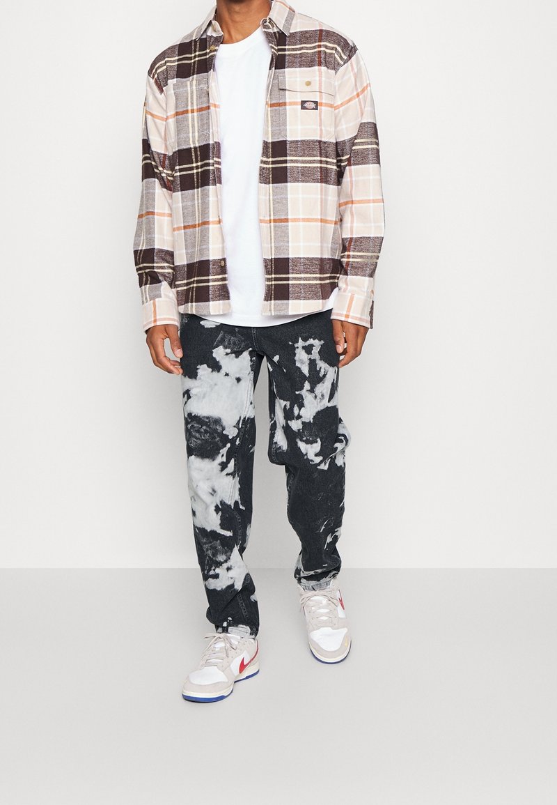 Man som bär en beige, brun och orange rutad skjorta över en vit t-shirt, svart och vit tie-dye jeans, samt vita sneakers med röd Nike swoosh.