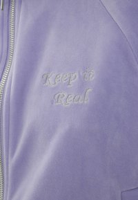 Lilas ziptröja med en texturerad yta. Har silverbroderad text "Keep it Real" på framsidan.