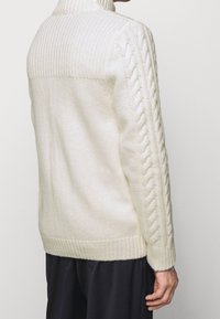 Krämfärgad stickad tröja med hög krage, ribbad textur och flätade detaljer på ärmar och sidor. Texturerat, mysigt tyg.