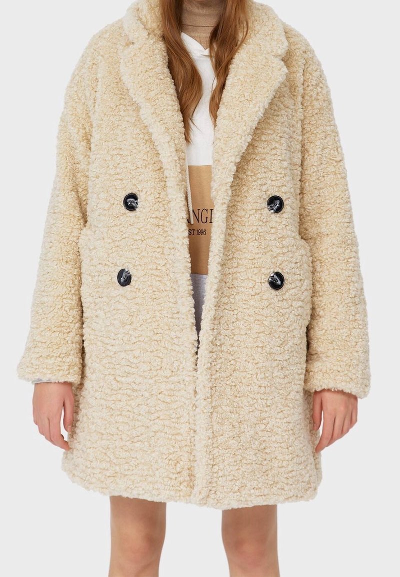 Beige, doppelt geknöpfter Teddy-Mantel mit großen schwarzen Knöpfen, getragen über einem weißen und beigen Kapuzenpullover, gezeigt von Hals bis zur Mitte des Oberschenkels.