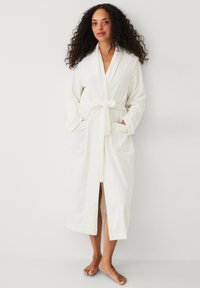 Vrouw met krullend haar, gekleed in een lange witte badjas met riem en zakken, staat blootsvoets tegen een effen lichte achtergrond.