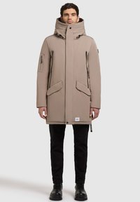 Beige parka takki, jossa on leveä kaulus, etuvetoketju, kaksi suurta sivutaskua ja yksi vetoketjullinen tasku hihassa; yhdistetty mustiin housuihin ja saappaisiin.