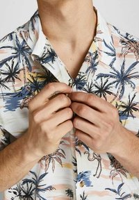 Camicia a maniche corte con colletto, di colore bianco, decorata con un motivo di palme e fenicotteri. Presenta dettagli colorati in tonalità pastello e bottoni marroni.