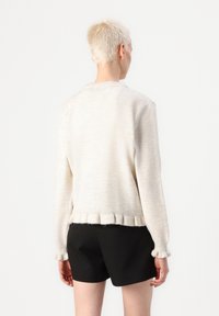 Cardigan color beige chiaro con una texture a coste, caratterizzato da un orlo arricciato e maniche lunghe, indossato sopra pantaloni corti neri.
