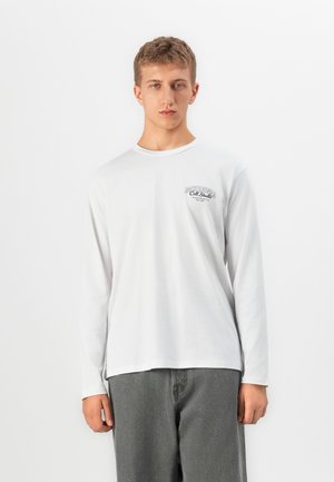 Young man with short blonde hair wearing a white long-sleeve shirt with small chest text and loose gray pants, standing against white background.