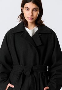 Čierny vlnený kabát so širokým golierom, opaskom v páse a zapínaním na gombíky. Má dlhé rukávy a predné vrecká, čo vytvára štruktúrovaný siluet.