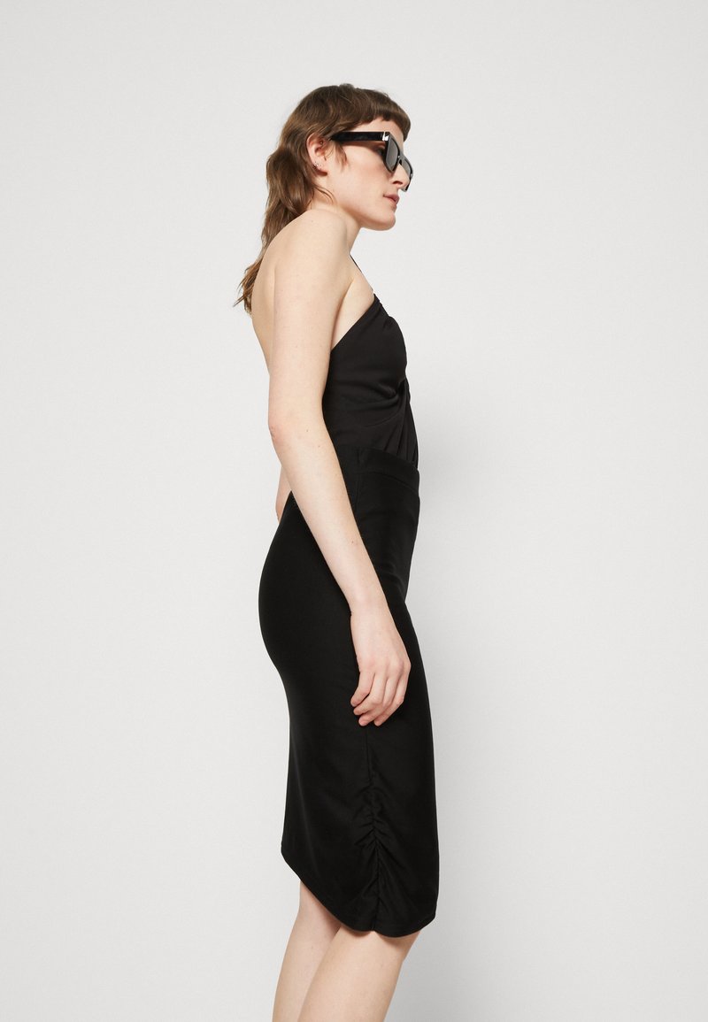 Schwarzes Midi-Kleid mit taillierter Passform, raffinierten Details am Saum und einer glatten Stofftexture. Kombiniert mit dunklen Sonnenbrillen.