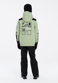 Grüne Jacke mit schwarzen Akzenten, grafisches Design auf der Rückseite mit Bergen und Sonne. Kombiniert mit schwarzen Hosen und Handschuhen.