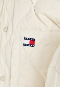 Giacca trapuntata color crema con chiusura a bottoni e una toppa logo sul petto sinistro in blu, rosso e bianco con la scritta "TOMMY JEANS."