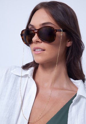 Tortoiseshell sunglasses with oversized frame, dark lenses, and gold chain looped through temples. Worn with a white button-up shirt.