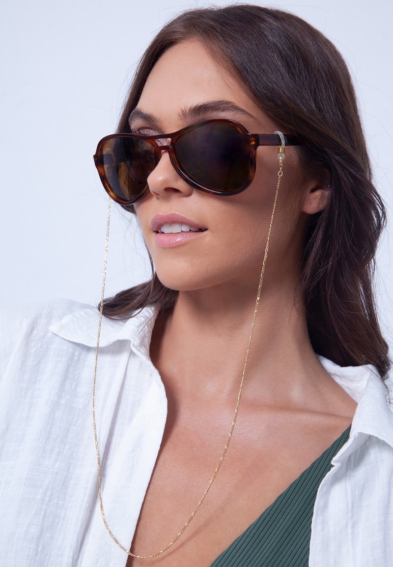 Occhiali da sole tartarugati con montatura oversize, lenti scure e catena dorata intrecciata tra le tempie. Indossati con una camicia bianca a maniche lunghe.