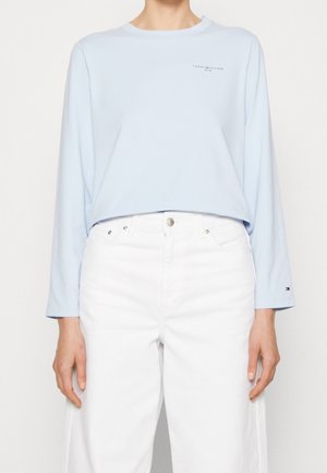 Chemise bleu clair à manches longues associée à un pantalon blanc taille haute, vue partielle des épaules aux hanches, sans détails en arrière-plan.