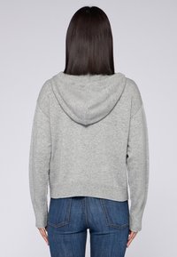 Jasnoszara krótka bluza z teksturowanym splotem, w oversize'owym kroju, z kapturem na sznurki. Na dole widoczne połączenie z dżinsami.