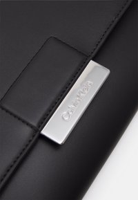 Černá kožená taška s elegantním designem, vyznačující se metalickým logem Calvin Klein, prošívanými okraji a hladkým povrchem.