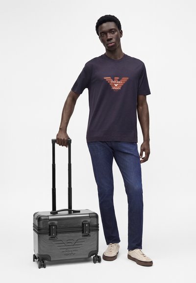 Άνδρας που φοράει σκούρο t-shirt Emporio Armani και τζιν, στέκεται κρατώντας τη λαβή μιας γκρι τροχήλατης βαλίτσας με τέσσερις τροχούς.
