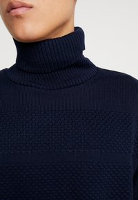 Maglione a collo alto in maglia blu navy con motivi a coste e strutturati. Presenta un colletto alto e un design leggermente aderente.