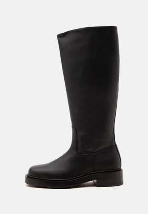 Grande botte en cuir noir avec un talon bas et un bout arrondi, vue de côté sur un fond clair uni.