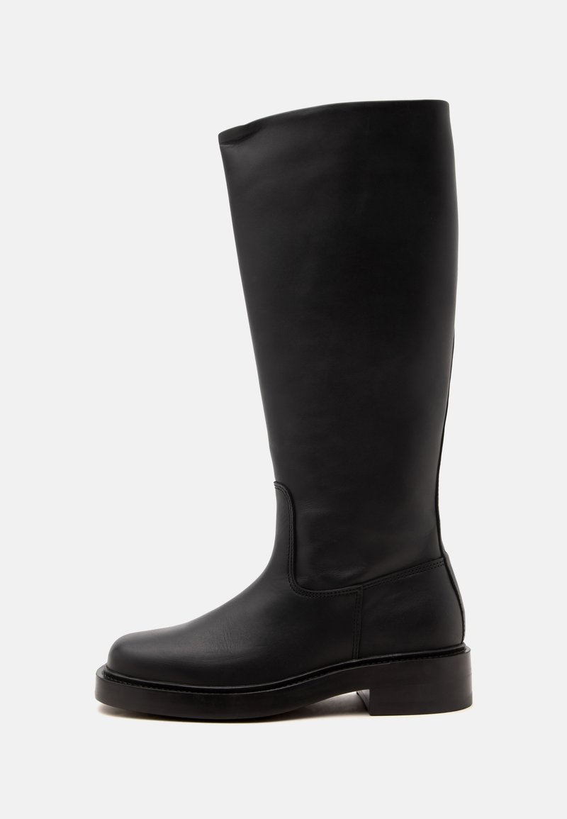 Grande botte en cuir noir avec un talon bas et un bout arrondi, vue de côté sur un fond clair uni.