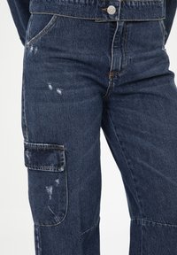 Pantaloni cargo in denim blu scuro con una texture consumata, dotati di una tasca laterale, cuciture visibili e chiusura con bottone in ottone.
