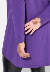 Längsarmiges lila Oberteil mit lockerer Passform, weichem Material und abgerundetem Saum, kombiniert mit schwarzen Leggings. Hand hinter dem Rücken positioniert.