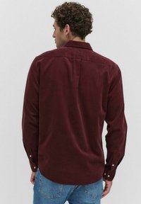 Man met krullend haar, gekleed in een donkerrode lange mouwen shirt en een blauwe spijkerbroek, van achteren gezien tegen een effen lichte achtergrond.
