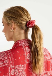 Chouchou en tissu rouge avec un motif cachemire blanc, tenant de longs cheveux bruns attachés en queue de cheval. Porté avec une chemise à motif rouge coordonnée.