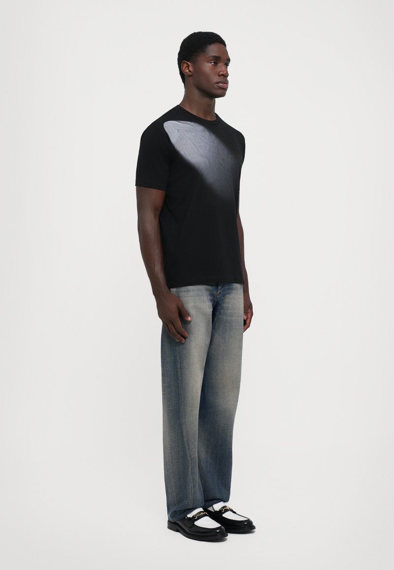Sort T-shirt med et gradient gråt mønster på den ene side, parret med lyseblå vidde jeans og sorte sko med hvide detaljer.